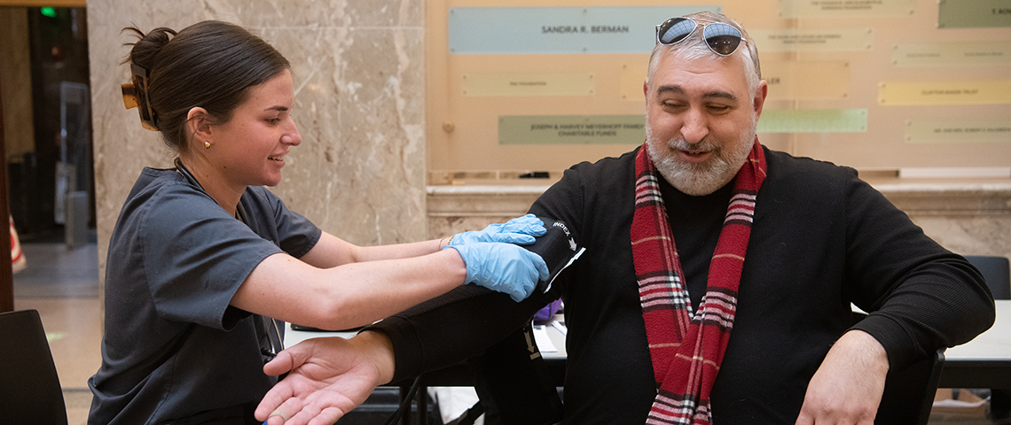 A white man gets his blood pressure checked by a female student in scrubs.