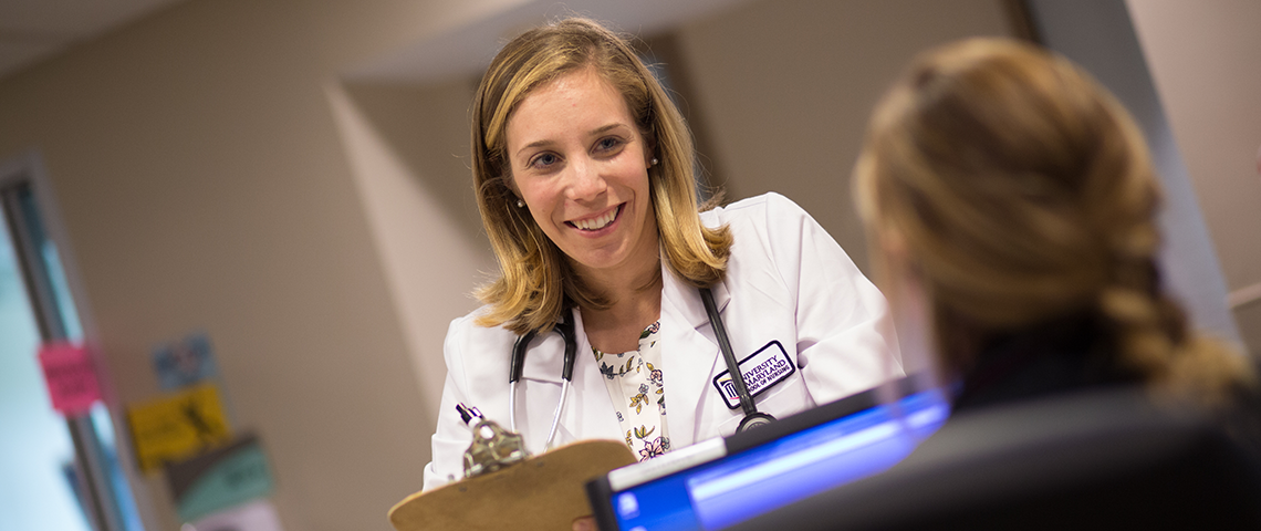 A woman in a white coat with a stethoscope chats with someone at a computer