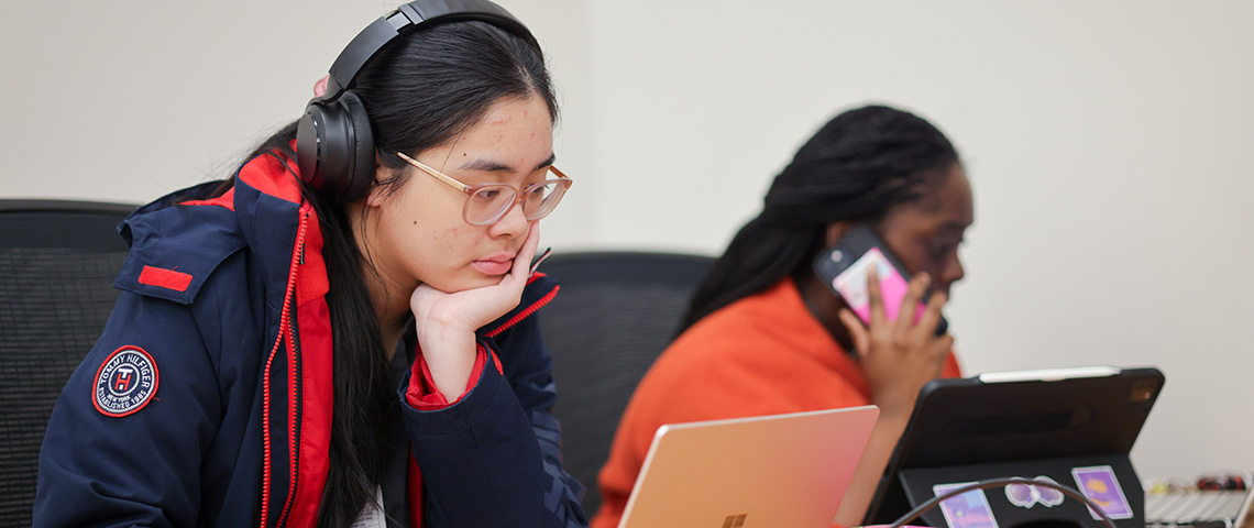 Two female students sit on laptops one is on her cell phone