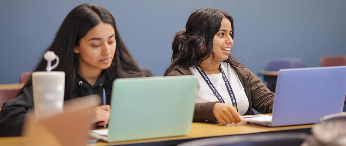 Two women sit in a class room with laptops in front of them