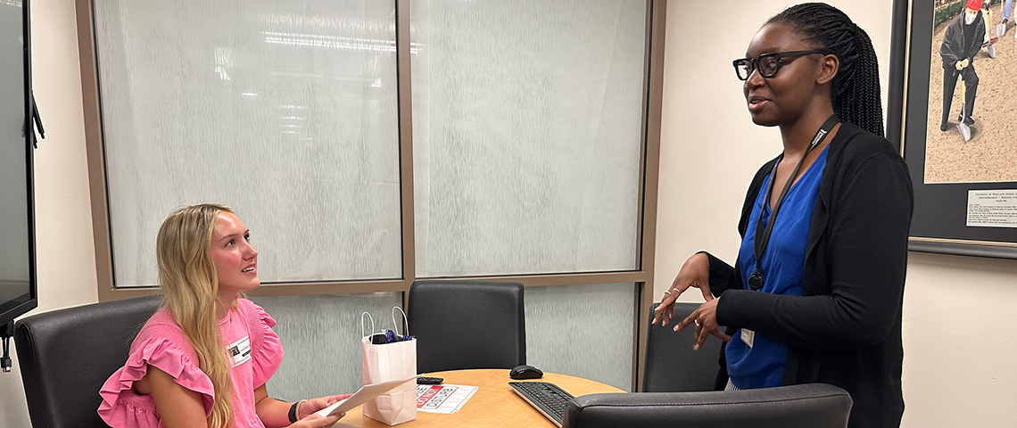 A woman on the left sits at a table looking up to the right at another woman standing across from her
