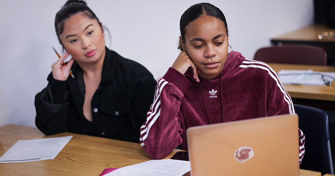 Two female students sit a desk, one is on her computer.