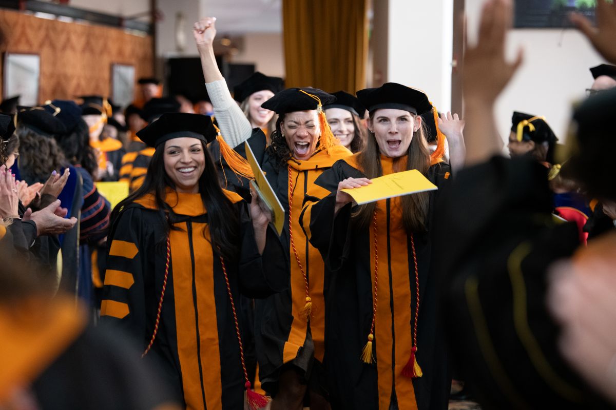 Graduates in black and orange gowns cheer joyfully as they walk down an aisle, celebrating at a commencement ceremony. Faces show excitement and triumph.