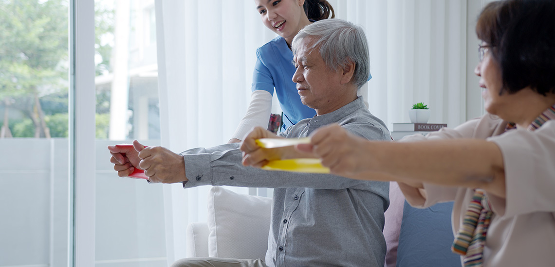 An elderly man and woman use exercise bands while a female nurse assists them