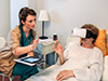 A nurse sits at the beside of an elderly woman wearing a virtual reality headset