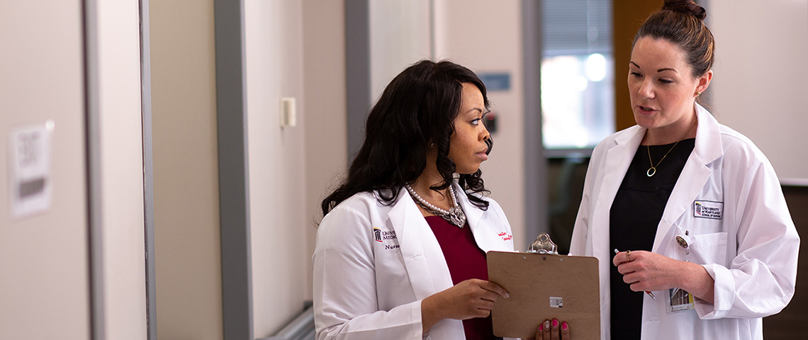 Woman in white coat with clipboard talks to another woman in a white coat in a hospital setting